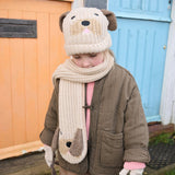 Kind draagt een beige gebreide Puppy-sjaal met hondengezichtje en een bijpassende muts bij een bruine jas.