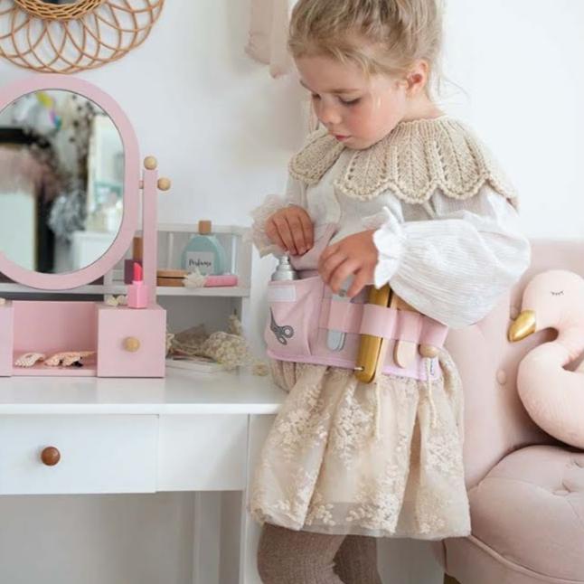 Kind draagt de roze kappersspeelset om het middel en speelt naast een kaptafel met spiegel.