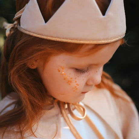 Gouden gezichtsglitter op de wang van een roodharig kind met een lichte kroon en cape.