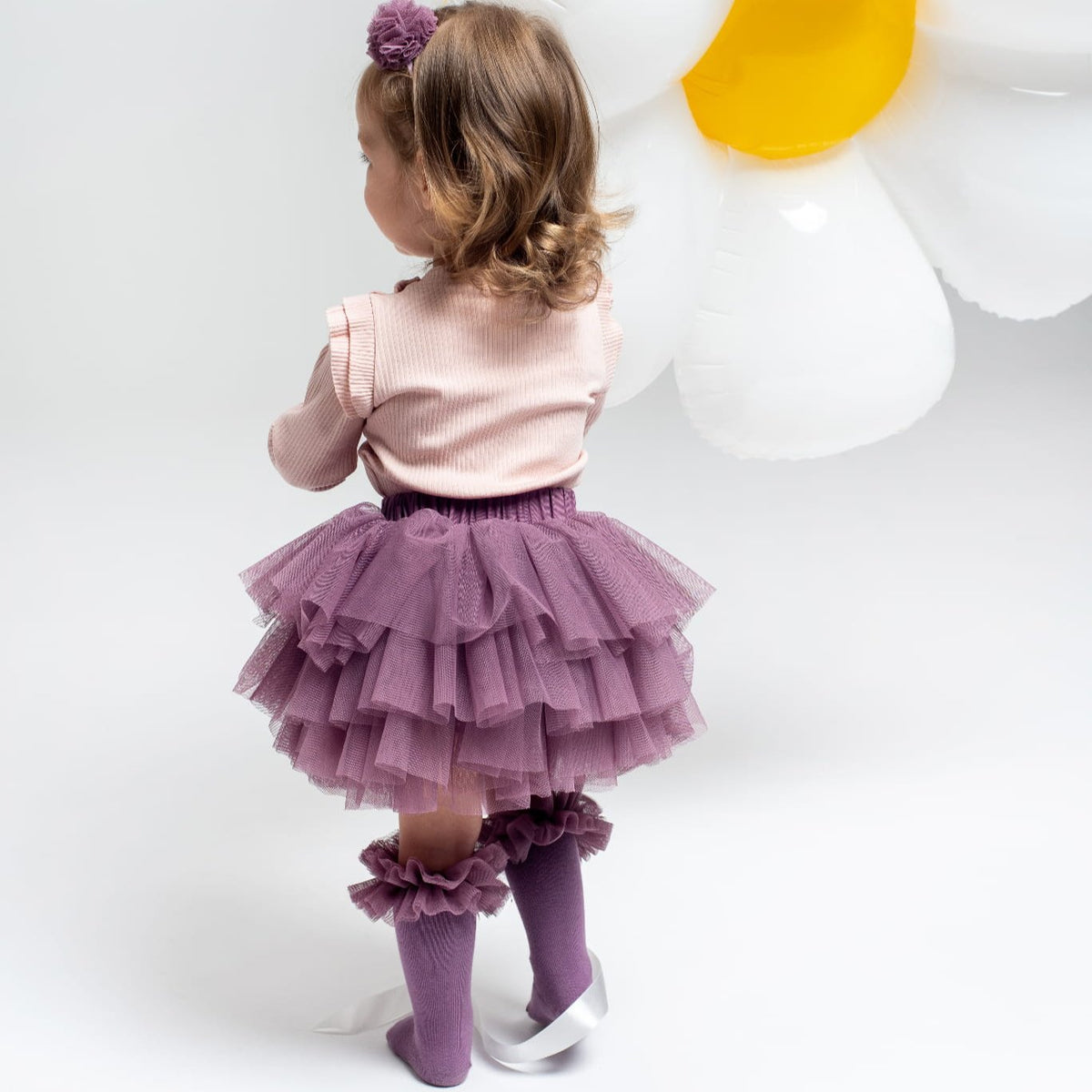 Kind van achteren met de oudspaarse Tutu Bloomer, een lichtroze top en paarse kniekousen met ruches.
