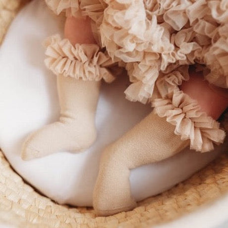 Baby met beige tutu kniekousen met ruches, liggend op een wit kussen in een gevlochten mand.