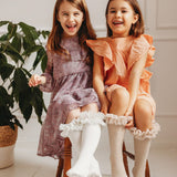 Twee lachende kinderen op een houten stoel, met witte en beige tutu kniekousen onder hun jurken.