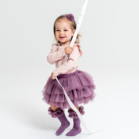 Kind met paarse tutu kniekousen met ruches, een paarse rok en een roze trui, met een wit lint in de hand.