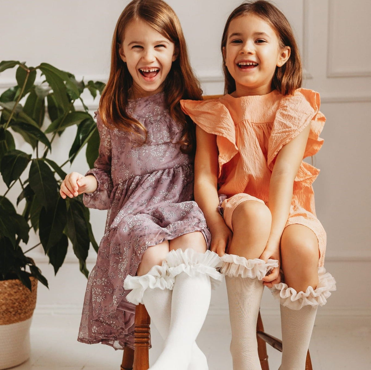 Twee lachende kinderen op een houten stoel, met tutu kniekousen onder een paarse en een perzikkleurige jurk.