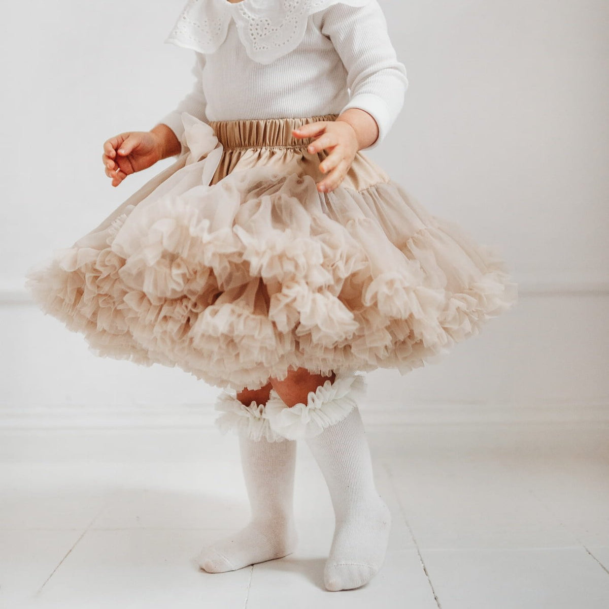 Kind met ecru tutu kniekousen met ruches, een beige rok met ruches en een witte top.