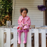 Kind met roze tutu kniekousen en een geruite jurk, zittend op een witte houten omheining.