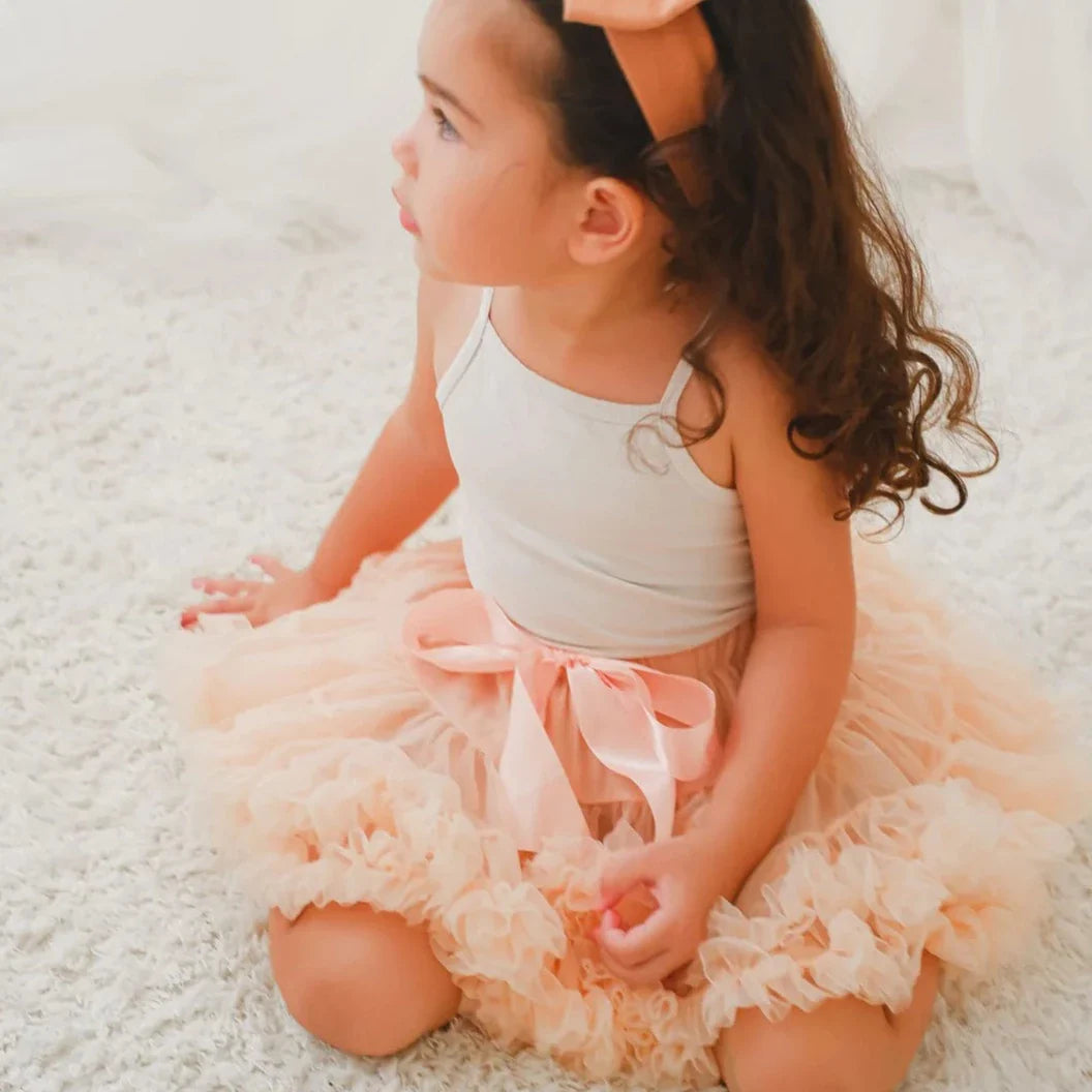 Meisje zit op een licht vloerkleed met de perzikkleurige Loli tutu rok met strik en ruches.