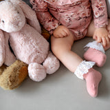 Baby met oudroze Vintage Love-kousen met witte kanten ruches, naast een roze knuffelkonijn.