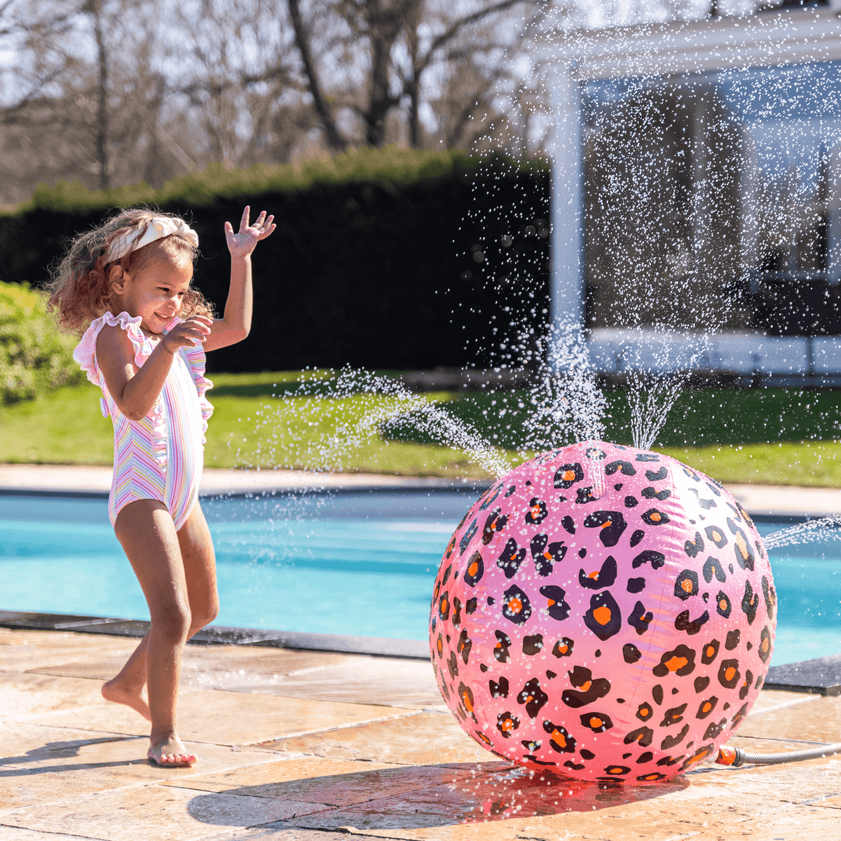 Kind speelt naast een roze watersproeier met panterprint die water spuit bij het zwembad.