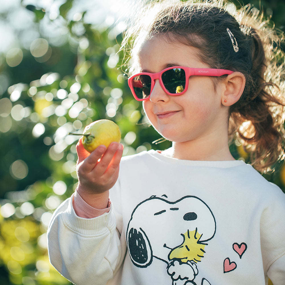 Kind met een roze Aspen zonnebril Camellia Rose houdt buiten een appel vast.