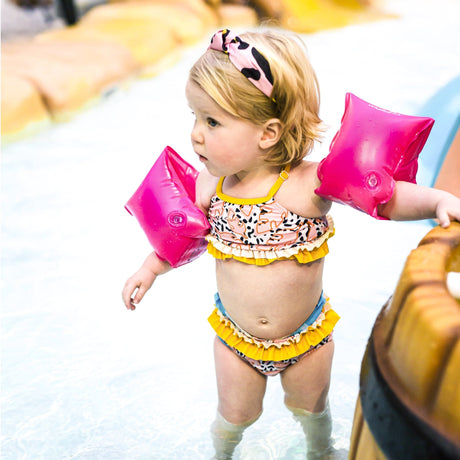 Kind met neonroze zwembandjes en een bikini met gele ruches in ondiep zwembadwater.