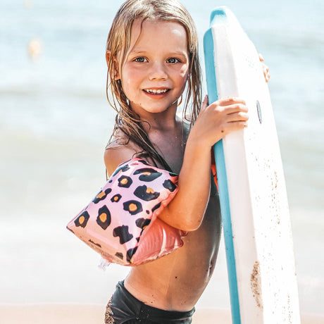 Meisje met roze zwembandjes met panterprint houdt een wit met blauw board vast op het strand.