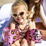 Meisje met zonnebril en roze zwembandjes met panterprint, zittend op het strand.
