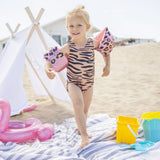 Meisje met roze zwembandjes met panterprint loopt over een strandlaken naast een witte speeltent.