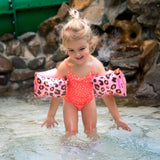 Meisje met roze panterprint zwembandjes en een koraalroze badpak speelt in ondiep water.