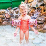 Meisje met roze panterprint zwembandjes staat met gespreide handen in het water voor een rotswand.